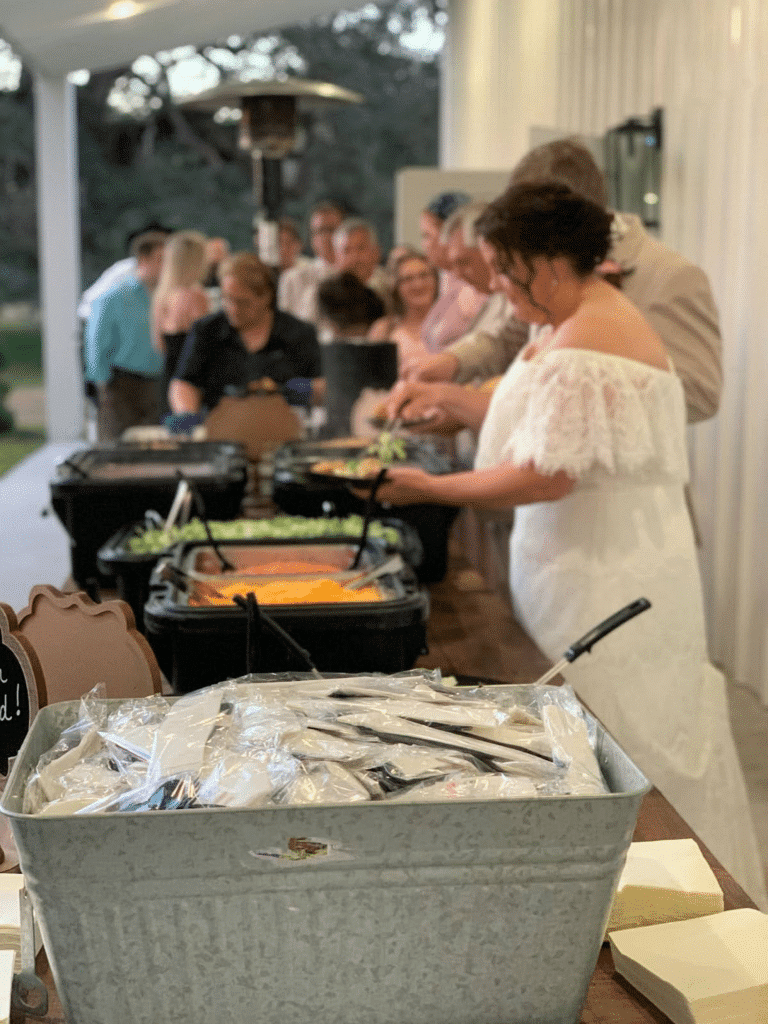 A bride and groom at a buffet-style wedding catering station with Tex-Mex dishes, including shredded cheese, lettuce, and salsa.