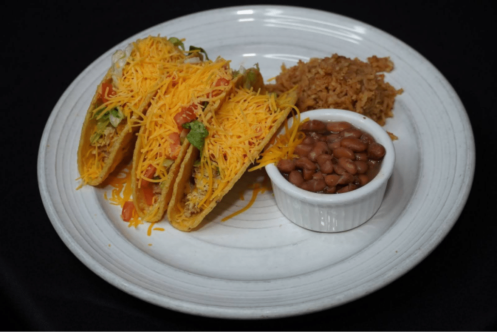 Three hard-shell tacos topped with shredded cheese, served with beans and rice.