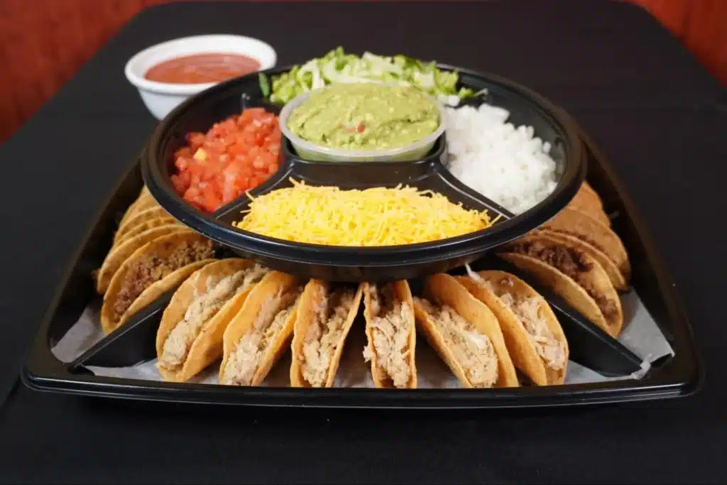 Taco platter with assorted beef and chicken tacos, served with sides of guacamole, shredded cheese, diced tomatoes, onions, and lettuce.