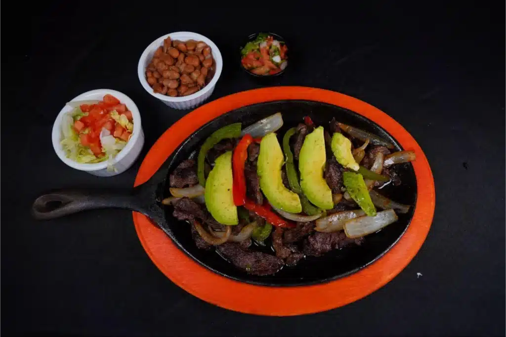 A cast-iron skillet filled with beef fajitas, grilled onions, bell peppers, and sliced avocado, served with sides of beans, pico de gallo, shredded lettuce, and diced tomatoes.