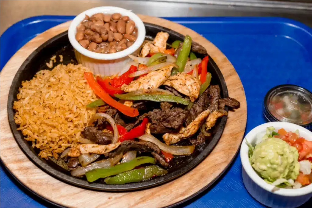 A sizzling platter of chicken and beef fajitas with sautéed bell peppers and onions, served with Mexican rice, a side of pinto beans, guacamole, pico de gallo, and salsa on a blue tray.
