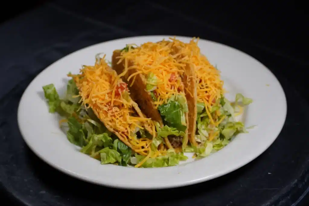 Three crispy tacos filled with beef, lettuce, and shredded cheddar cheese on a white plate.